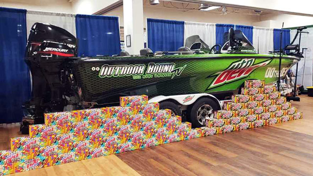 Outdoor Bound TV and Mountain Dew branded boat on a trailer behind a display of Mountain Dew Spark 12-pack product boxes