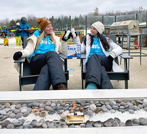 two women siting in chairs outside looking toward each drinking cans of Bubblr in front of a fire in the winter