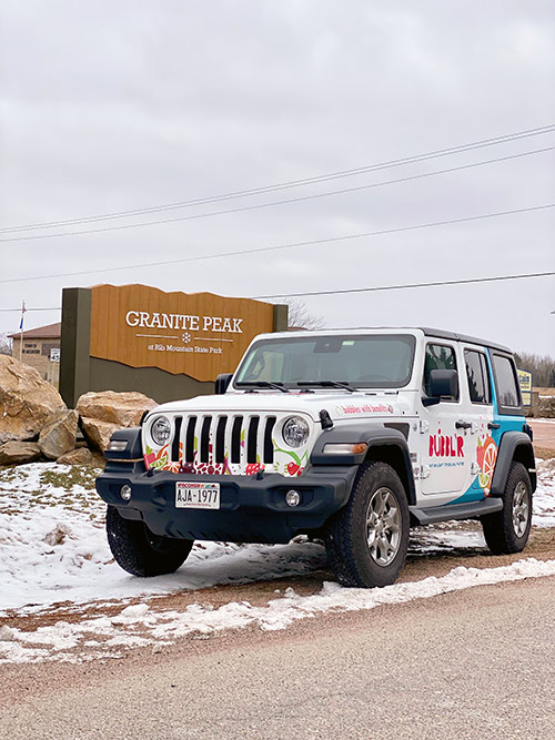 a white Jeep with Bubblr wrap parked in front of the Granite Peak road sign during winter