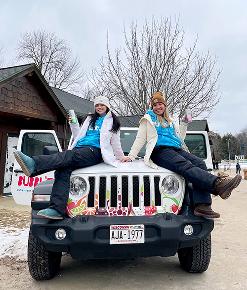 two women in winter clothes posing on top of the hood of a white Jeep with a Bubblr wrap holding cans of Bubblr