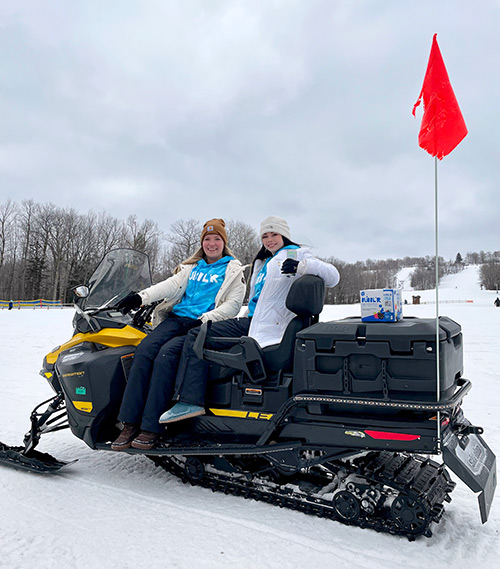 two women sitting on a snowmobile outdoors with a case of Bubblr sitting on the back