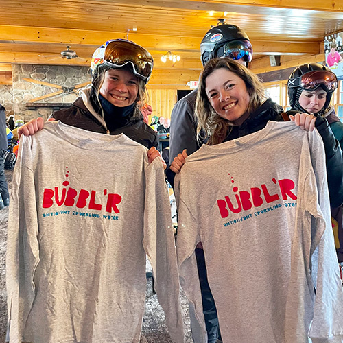 two woman inside of a ski lodge holding up Bubblr shirts and smiling