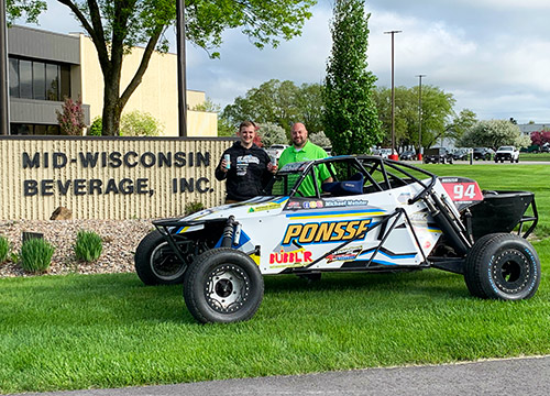 two men smiling and posing with cans of Bubblr in their right hand standing behind a parked dune buggy with the name Ponsse on the side parked on the grass in front of the Mid-Wisconsin Beverage, Inc. headquarters sign