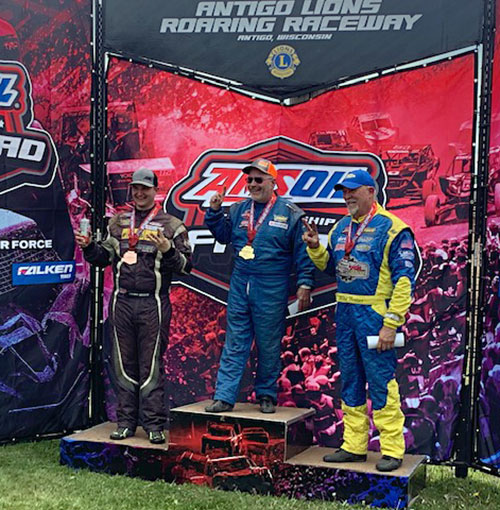 three racecar drivers standing on a three-level podium with first, second, and third places medals at the Antigo Lions Roaring Raceway awards ceremony