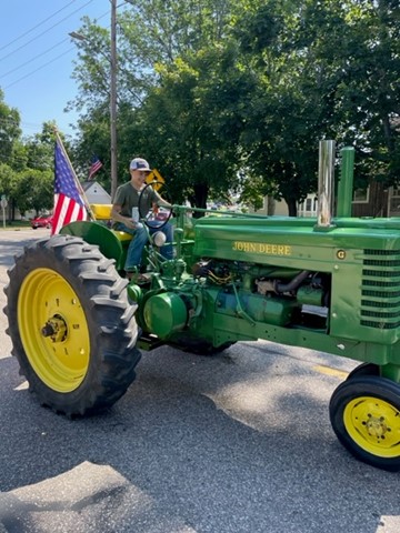 a person holding a soda can rides a green tractor with yellow wheels and an American flag