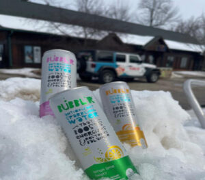 MG-Bubblr-Event-cans-Jeep three cans of Bubbl'r sit in a snowbank while Bubbl'r Jeep is visible in the background