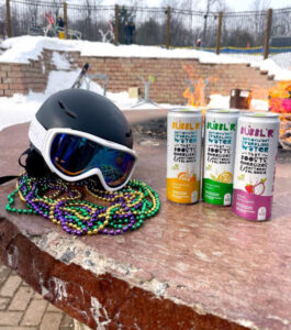 MG-Bubblr-Event-helmet-beads-cans three cans of Bubbl'r sit next to a snowboard helmet with green, yellow, and purple Mardi Gras beads