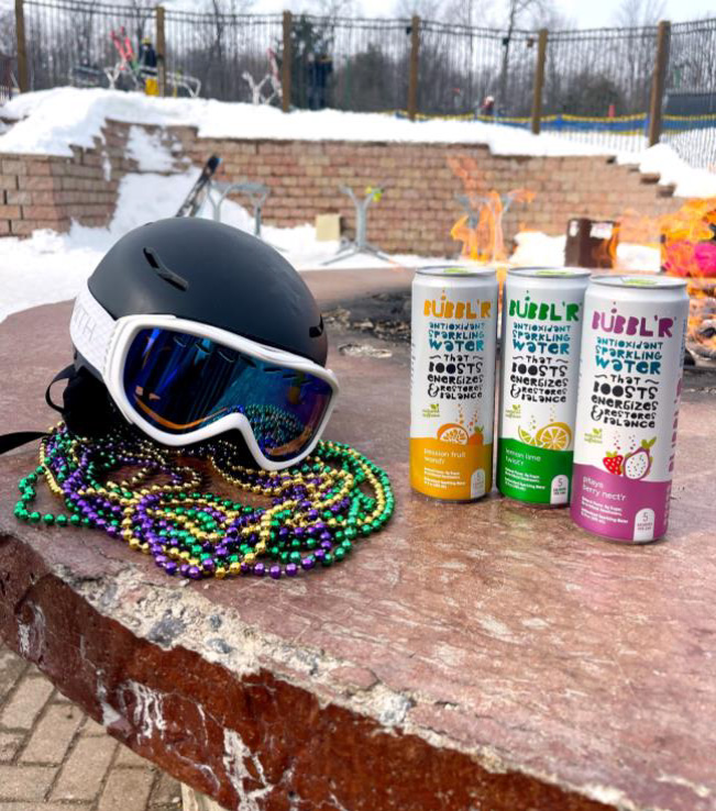 three cans of Bubbl'r sit next to a snowboard helmet with green, yellow, and purple Mardi Gras beads