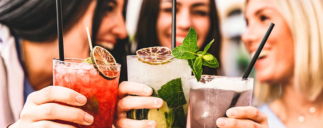 three young women clinking glasses with colorful mixed drinks in them